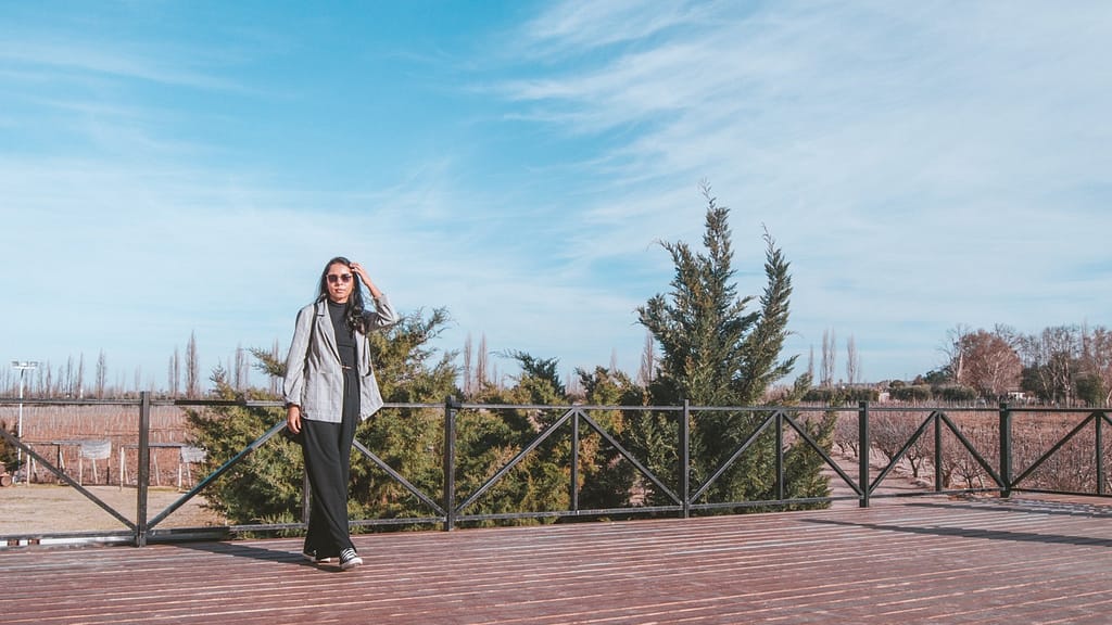 A visitor stands on a wooden deck with a serene expression, surrounded by the natural beauty of Mendoza's wineries, with evergreen trees and a clear sky, capturing the essence of a tranquil escape in Argentina's premier wine country.