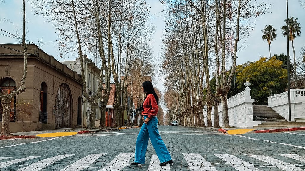 A woman with long hair, wearing blue jeans, boots, and a red hoodie, crossing the pedestrian crosswalk in the old town of Colonia del Sacramento. With colonial houses in the background and a street lined with leafless trees due to winter, fading into the horizon.