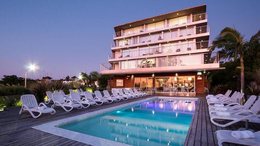 Twilight view of Costa Colonia Riverside Boutique Hotel in Colonia del Sacramento, with its modern multi-story structure overlooking a tranquil swimming pool lined with white lounge chairs, illuminated by soft poolside lighting.