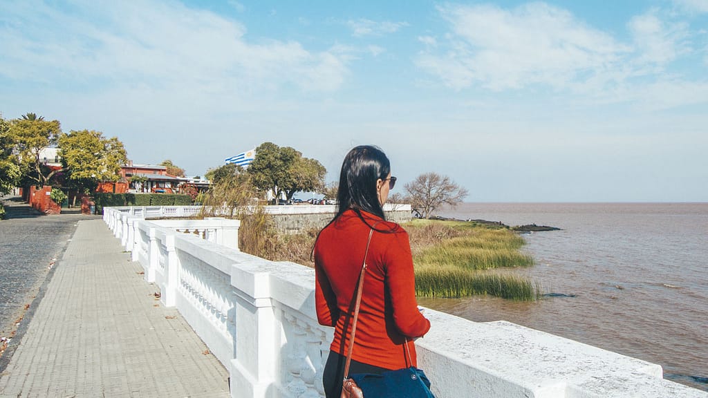 A woman in a red sweater and jeans stands facing the Rio de la Plata at the Bastión de San Pedro in Colonia del Sacramento, leaning on a white balustrade, with views of the coastline and Uruguayan flag in the distance.