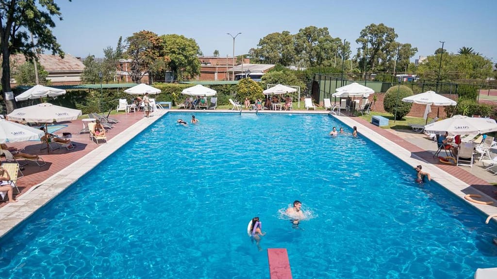 Sunny day at El Mirador Hotel & SPA in Colonia del Sacramento featuring guests enjoying the large outdoor swimming pool, surrounded by sun loungers, parasols, and lush greenery.