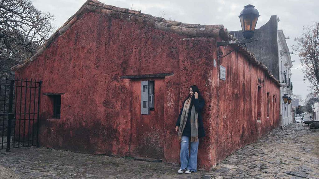 A person standing next to the weathered red facade of the Vivienda Portuguesa in Colonia del Sacramento, with its rustic colonial architecture, cobbled street, and traditional lanterns creating an atmosphere of historical charm.