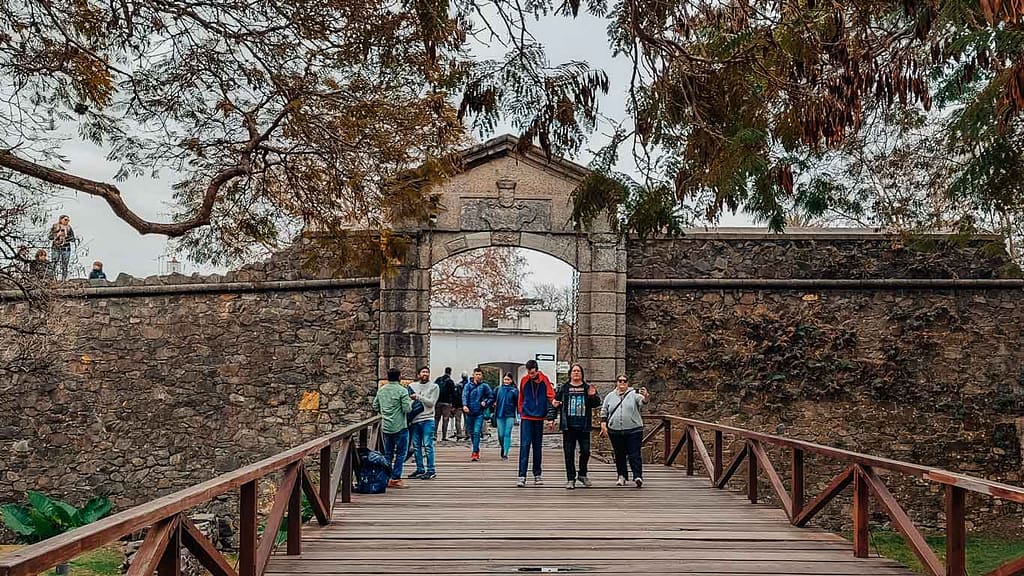 Visitors walking across a wooden bridge towards the Portón de Campo, the historic city gate of Colonia del Sacramento, with its distinctive stone archway and surrounding weathered walls, framed by lush trees.