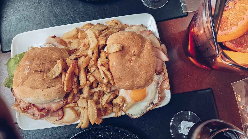 A classic Uruguayan chivito sandwich with a sunny-side-up egg on top and a side of fries on a white plate, with a glass of sangria in the backdrop, indicative of a meal at a Colonia del Sacramento restaurant.