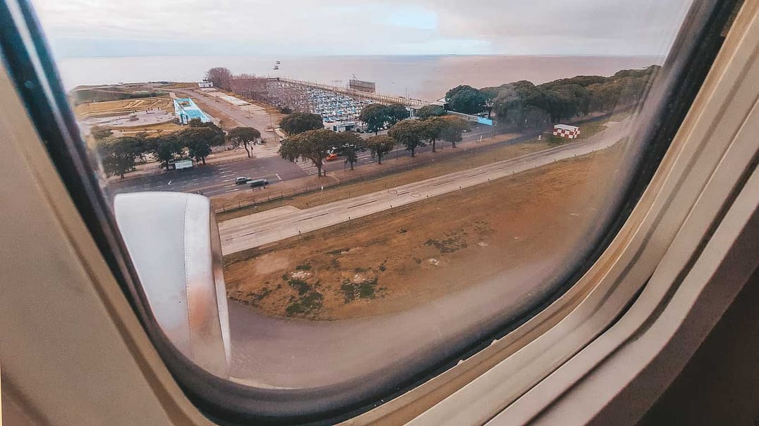 The image depicts a view from an airplane window looking out over a coastal urban landscape during daylight. Visible through the window are the plane's wing and engine, a broad park-like area with geometric walkways and trees, a packed marina, and a distant horizon where the sky meets the sea. The scene conveys the moment of either departure or approach over a cityscape.