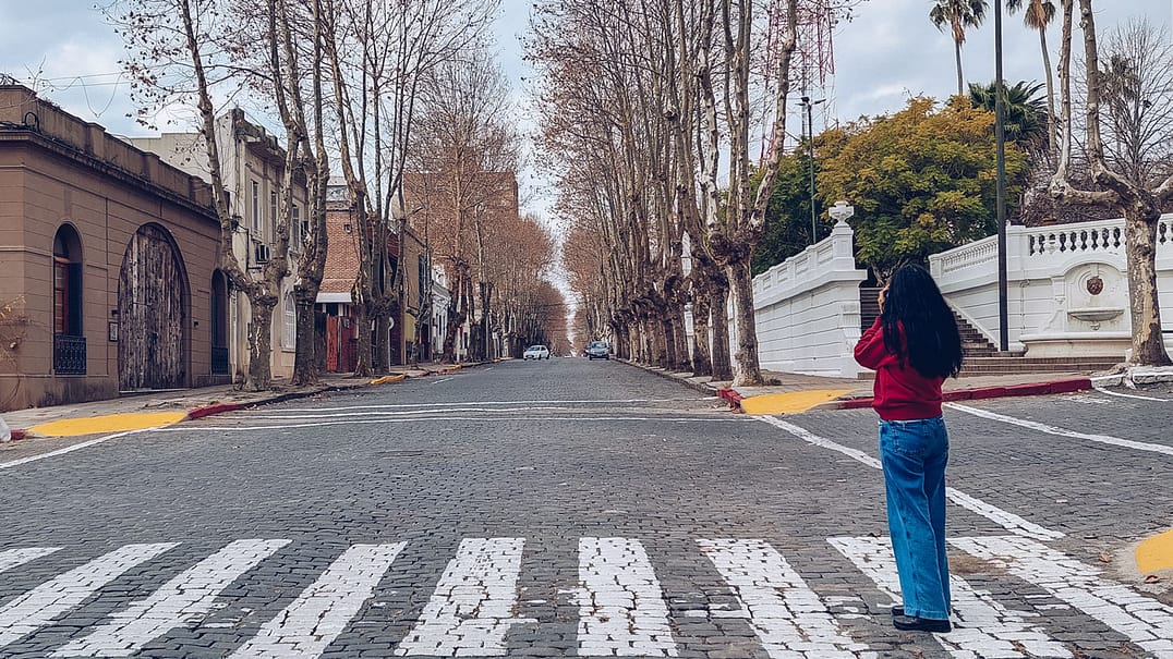 A woman stands on the pedestrian crossing, taking a photo with her camera on a street in Colonia del Sacramento filled with colonial houses. She has long black hair and is wearing blue jeans, boots, and a red sweatshirt.