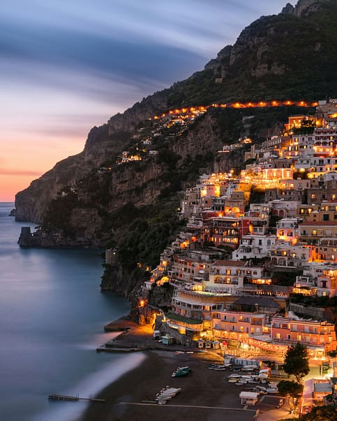 Evening view of Positano, Italy, with its terraced, multicolored buildings aglow with lights along the cliffside, overlooking a calm sea.