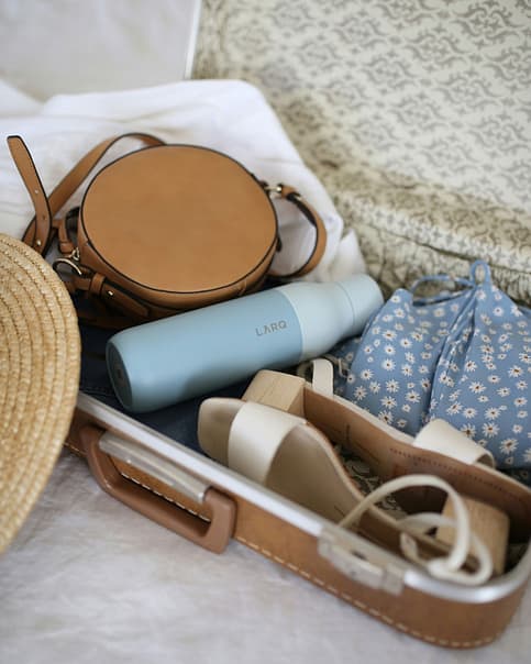 Open suitcase with summer travel essentials for Europe, including a stylish tan leather crossbody bag, a blue floral drawstring pouch, beige sandals, a LARQ water bottle, and a straw hat, encapsulating a Packing Checklist for Europe in Summer.