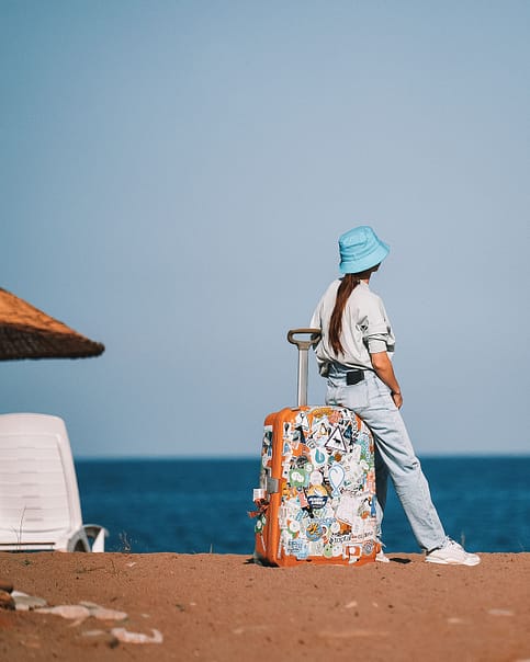 A traveler stands beside a brightly sticker-adorned suitcase on a sandy beach, gazing out at the sea. This image evokes the spirit of summer in Europe.