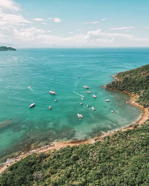 Aerial view of a serene bay in Buzios, Brazil, showcasing a curve of sandy shoreline with lush greenery, surrounded by the turquoise waters of the Atlantic Ocean dotted with various boats and yachts.