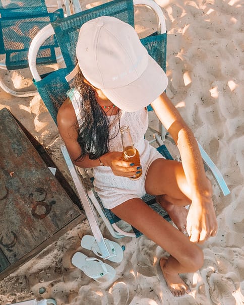 Relaxed beach vibes captured with a person lounging on a blue beach chair, holding a bottle of beer, complemented by white flip-flops on the sandy beach, embodying the laid-back atmosphere of Barra da Tijuca in Río de Janeiro.
