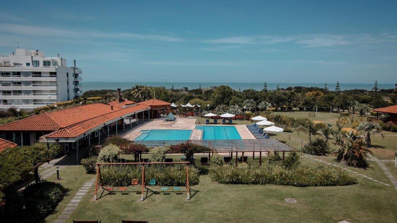 This image captures a scenic view of the IL Belvedere Hotel in Punta del Este. The hotel's layout includes a sprawling pool area surrounded by umbrella-covered lounge chairs, a series of red-tiled roof bungalows, and lush, well-maintained gardens. In the backdrop, you can see a modern, high-rise building and the serene coastline, emphasizing the hotel's prime location between natural beauty and urban convenience.