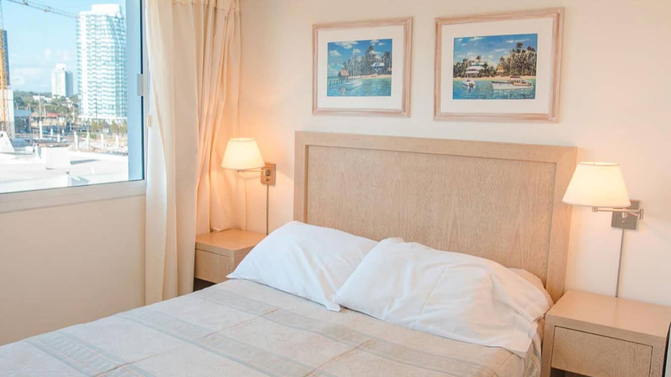 This image features a cozy hotel room at Sunset Beach in Punta del Este. The room is equipped with a comfortable bed with a beige, textured headboard, complemented by soft white bedding. Natural light filters through the window revealing a view of the urban landscape and harbor. The walls are adorned with framed paintings depicting scenic landscapes, which add a touch of elegance and a connection to local sights.