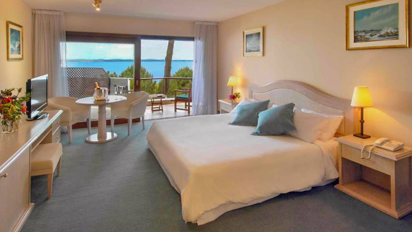 This image features a well-appointed room at the Hotel del Lago Golf & Art Resort in Punta del Este. The room is tastefully decorated with a large, comfortable bed, light pastel walls, and framed artwork that complements the serene ambiance. A small dining area by the balcony provides a perfect spot for a meal or relaxation, offering stunning views of the blue waters through floor-to-ceiling glass doors. The overall setting conveys a peaceful, inviting atmosphere, ideal for a tranquil retreat by the lake.