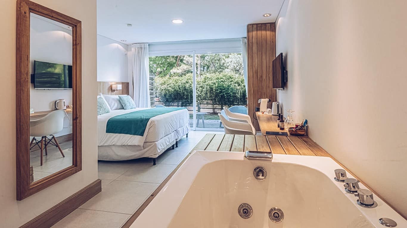 This image showcases a luxurious guest room at Live Hotel Boutique in Punta del Este. The room features a spacious layout with a large bed topped with a teal bedspread, creating a soothing aesthetic. The room opens up to a private patio through full-length sliding glass doors, bringing in ample natural light and offering a view of lush greenery. Additionally, a large whirlpool bathtub is prominently positioned in the room, adding a touch of indulgence. The room's design is modern and minimalist, emphasizing comfort and elegance.