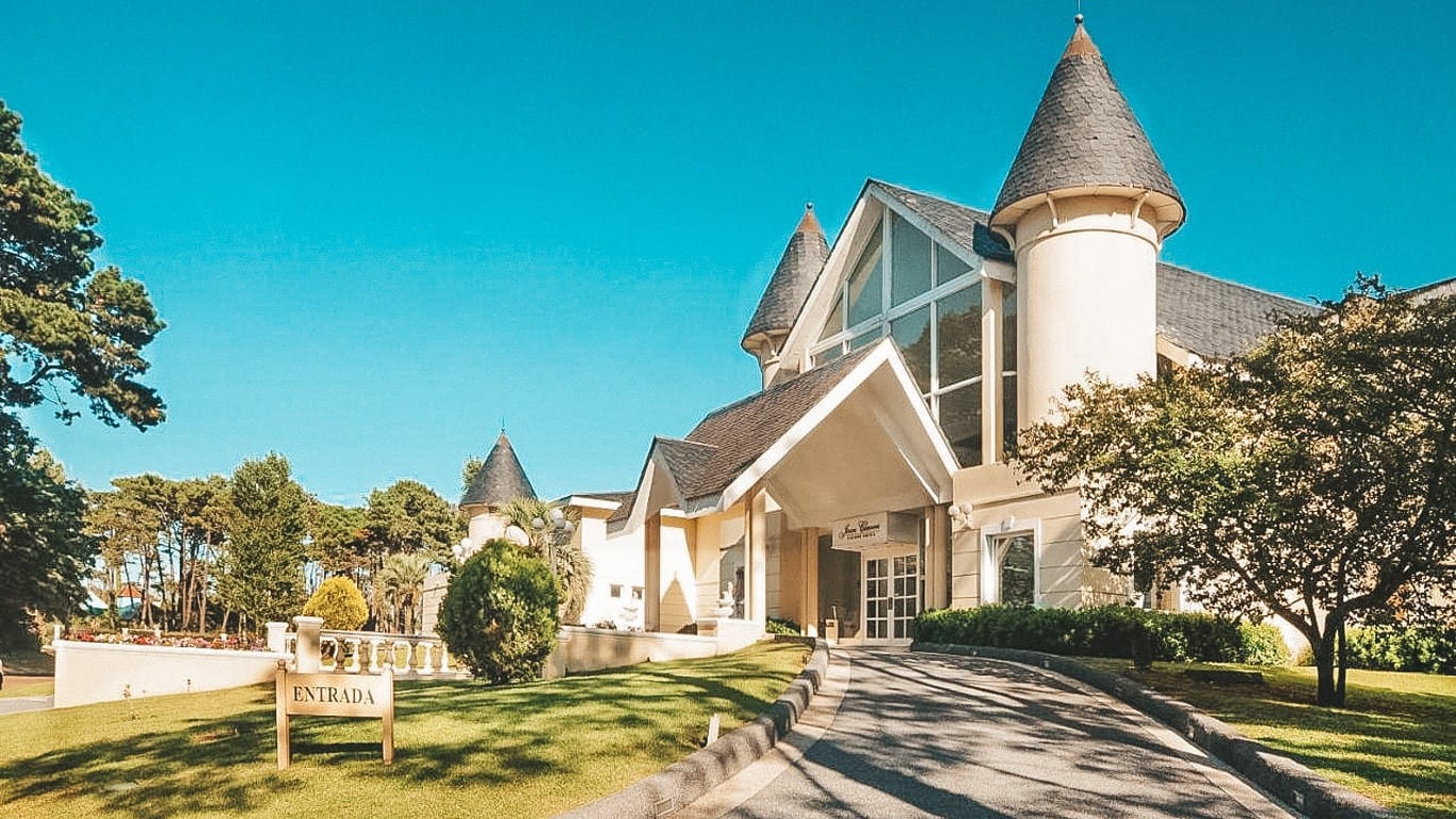 This image features the grand facade of the Parque Hotel Jean Clevers in Punta del Este. The hotel is designed in an elegant, fairy-tale style with pointed towers and steep roof lines. The entrance is marked by large, clear glass windows and a spacious driveway leading to a modern, welcoming entrance. The lush greenery surrounding the building and the clear blue sky enhance the picturesque setting, offering a tranquil and luxurious ambiance.