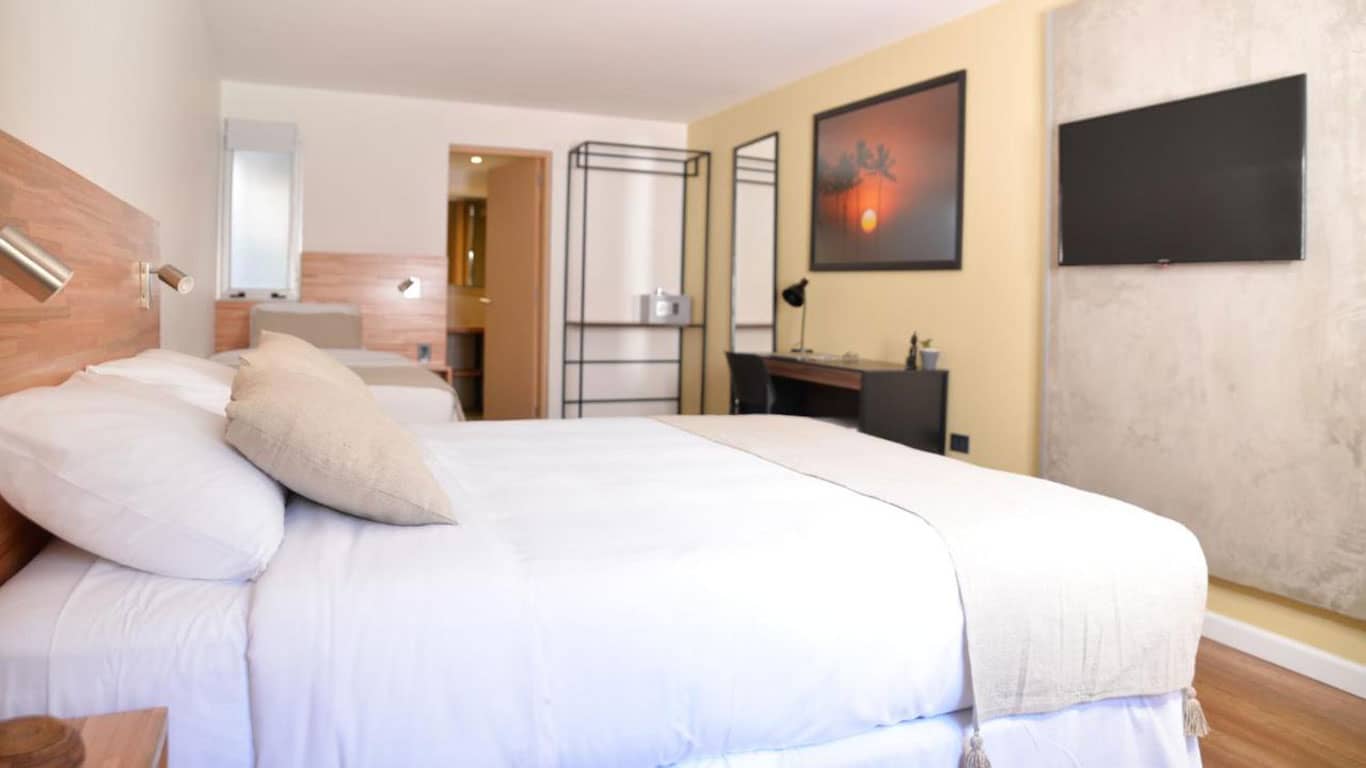 A modern hotel room at TAS D VIAJE Suites in Punta del Este featuring a large bed with plush white bedding and beige pillows. The room is equipped with a flat-screen TV on the wall, a small desk with a chair, and minimalist wooden decor. There's an open doorway leading to another room, adding depth and spaciousness to the setting.