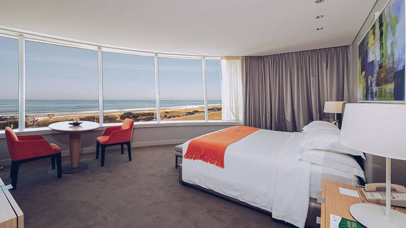 This image features an elegant hotel room at The Grand Hotel in Punta del Este. The room is designed with large panoramic windows that offer a breathtaking view of the sandy beach and ocean. Furnished with a comfortable bed, two red chairs, and a small round table, the room’s decor is complemented by a contemporary painting and soft, neutral colors, creating a relaxing atmosphere that enhances the scenic seaside view.