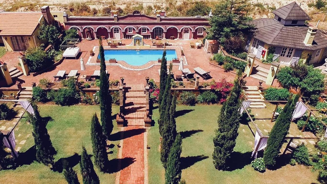 This aerial image captures the luxurious outdoor setup of Villa Toscana Boutique Hotel in Punta del Este. The hotel features a classical Mediterranean architectural style with a spacious courtyard centered around a large, inviting swimming pool. Surrounding the pool are various lounging areas, garden paths, and meticulously maintained lawns lined with tall, slender cypress trees, enhancing the villa’s Tuscan-inspired ambiance. 