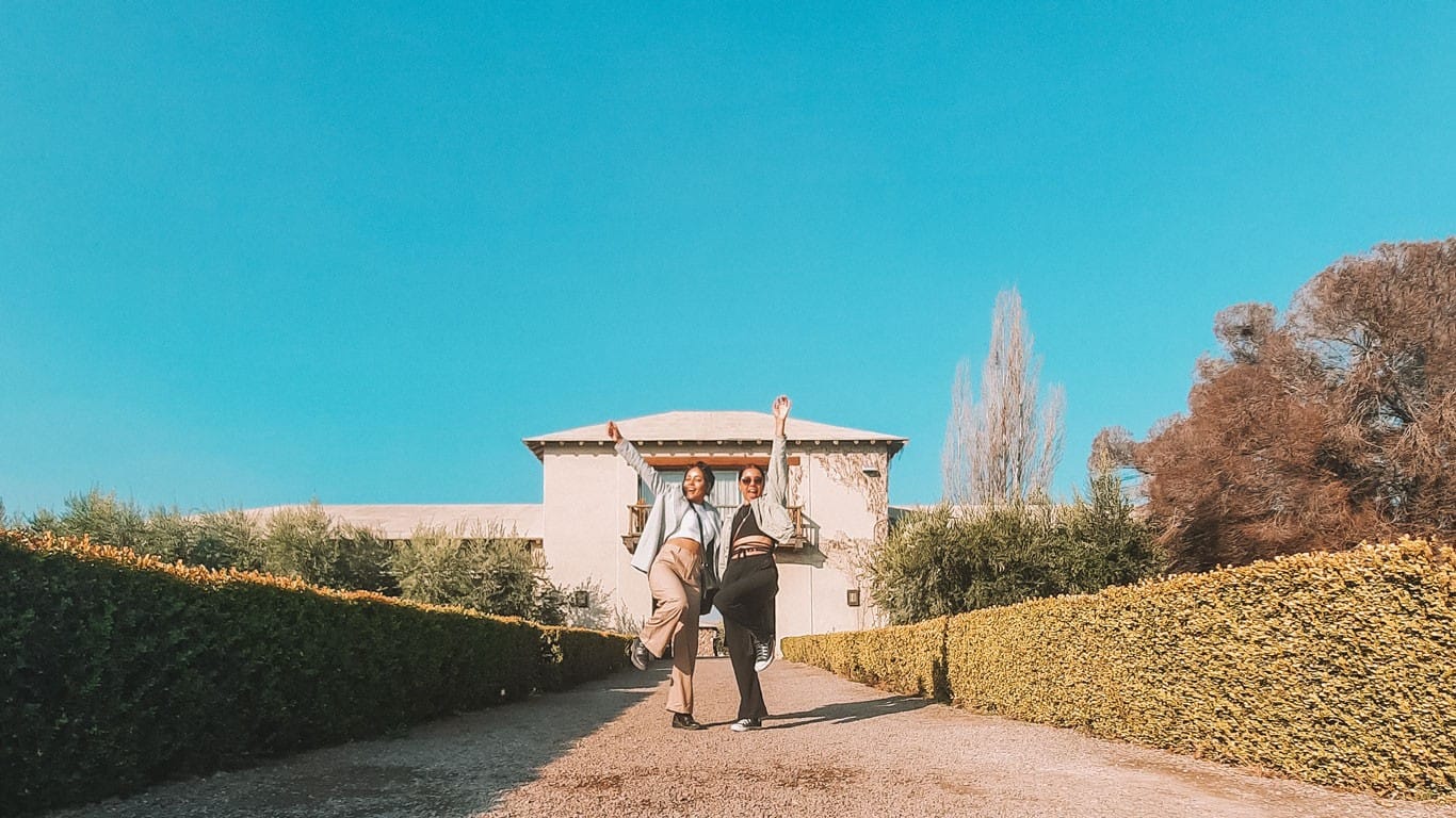 Two joyful visitors strike a playful pose on a gravel pathway, with the rustic charm of Bodega VistaAlba in Lujan de Cuyo, Mendoza, behind them. They are framed by well-manicured hedges under a clear blue sky, encapsulating the essence of a lively visit to this renowned wine estate