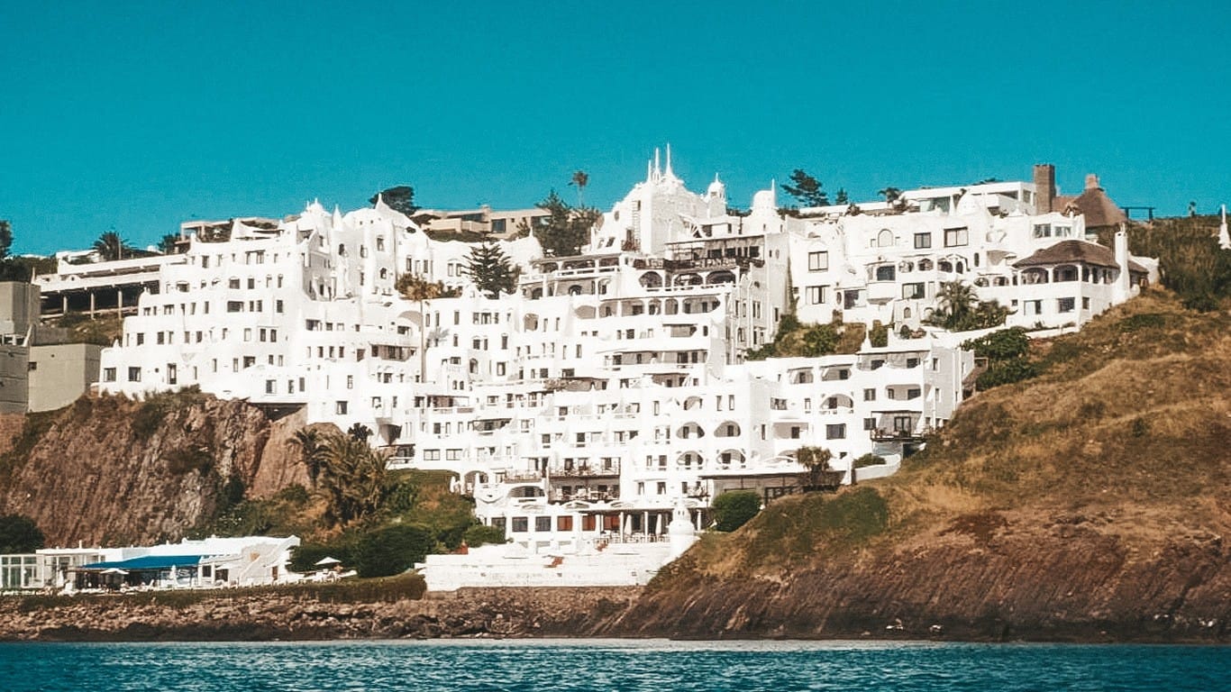 This image showcases the iconic Club Hotel Casapueblo in Punta del Este, Uruguay. Perched on a rocky cliff overlooking the ocean, this hotel is distinguished by its unique, sprawling architecture with cascading terraces and white-washed walls, reminiscent of a sculpture. The building's organic, irregular design stands out against the natural rugged coastline, making it a visually captivating landmark.