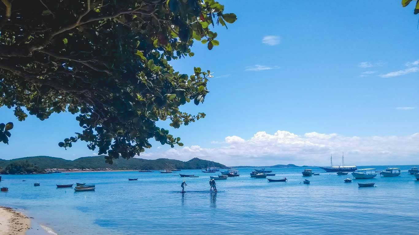View of Buzios, Brazil, from under the shade of a tree, capturing the tranquil azure waters peppered with anchored boats and a distant view of the rolling landscape and cloud-speckled blue sky.