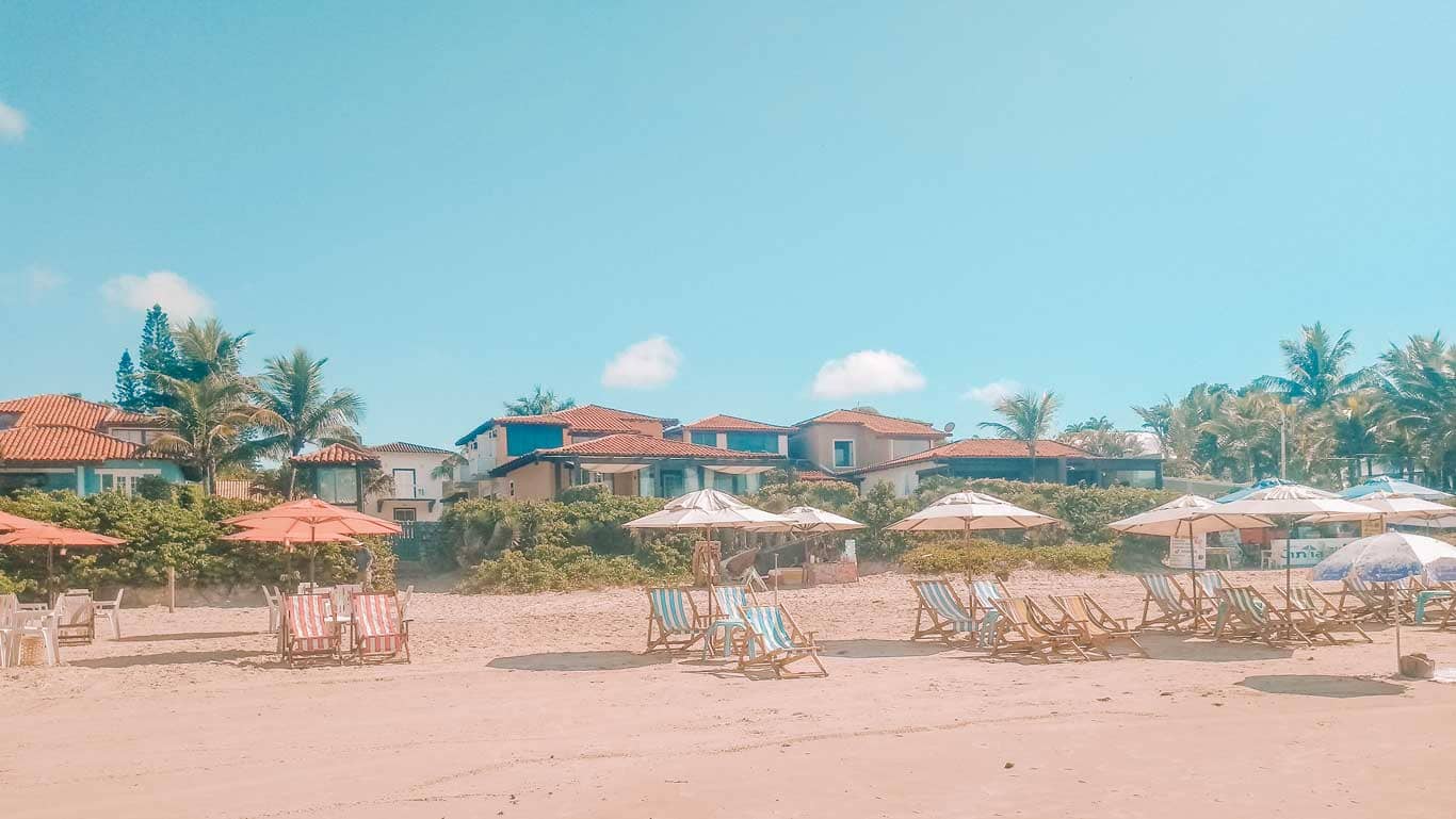 Sunny day at Geribá Beach with rows of inviting beach chairs and open umbrellas on the sand, fronting charming houses with red-tiled roofs and lush palm trees, capturing the essence of a relaxing getaway in Buzios, Brazil.