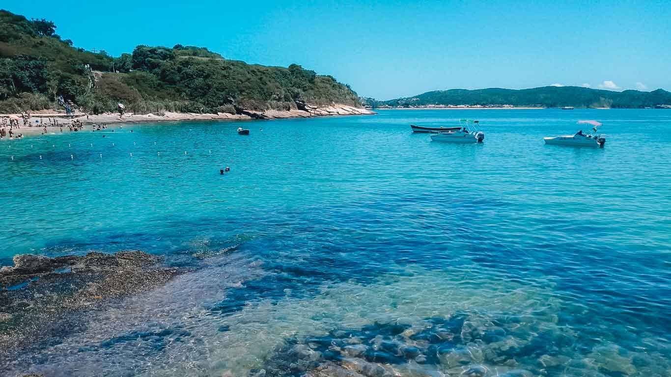 Crystal-clear waters of Joao Fernandes beach in Búzios, a prime location for travelers pondering where to stay in Búzios, featuring gently rocking boats and swimmers enjoying the tranquil sea, with a picturesque hillside in the background.