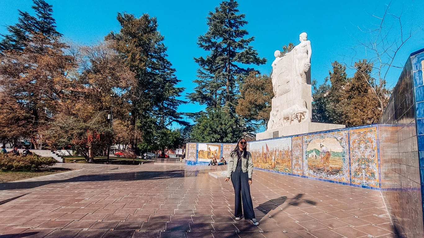 A traveler stands on a wide promenade in Mendoza city center, with a striking statue and colorful tiled murals in the background, capturing the artistic and cultural atmosphere of a sunny day in this central Argentinian locale.