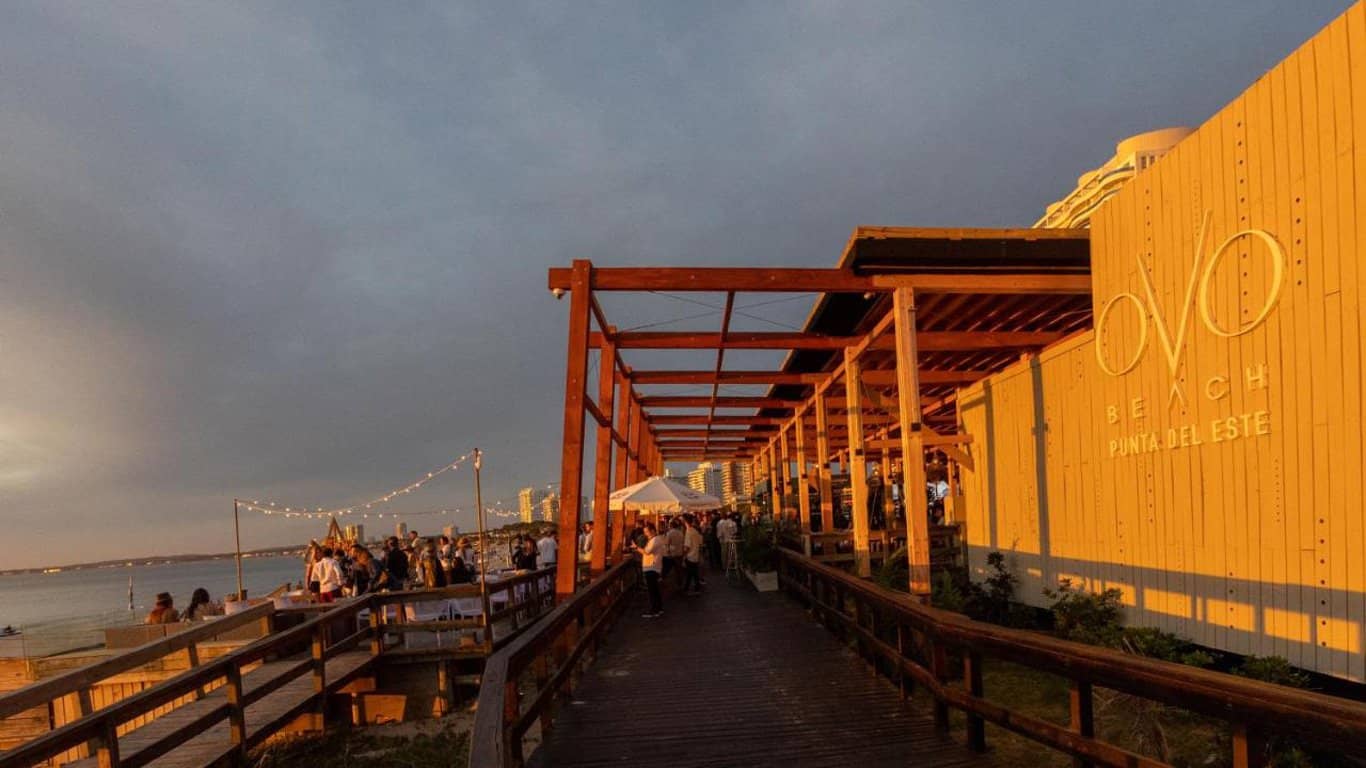 This image showcases a bustling beachside venue in Punta del Este during sunset. People are mingling on a wooden boardwalk that leads to a venue branded "OVO Beach." The venue's architecture features a modern, minimalist design with a large "OVO" sign illuminated in white light against a wooden facade. String lights add a cozy ambiance above the boardwalk, and the skyline in the background is bathed in the golden hues of the setting sun.