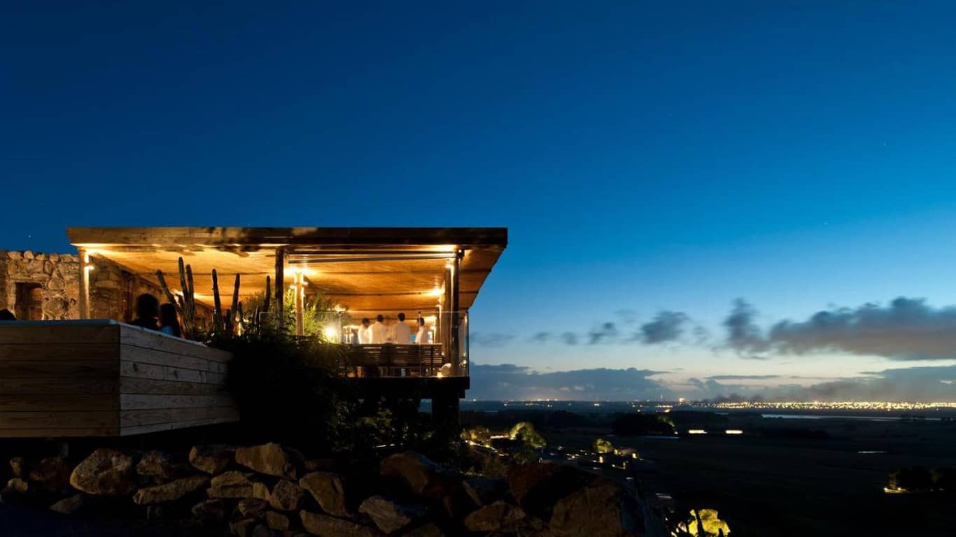 This image captures a serene evening at Hotel Fasano in Punta del Este. The scene is set on a stylish terrace perched atop a rocky hill, with people gathered around the warmly-lit bar area. The terrace offers a panoramic view of the surrounding landscape, lit by a sprawling network of city lights under a twilight sky. The natural stone and wood architecture, along with scattered cacti, add a rustic charm to the modern ambiance.