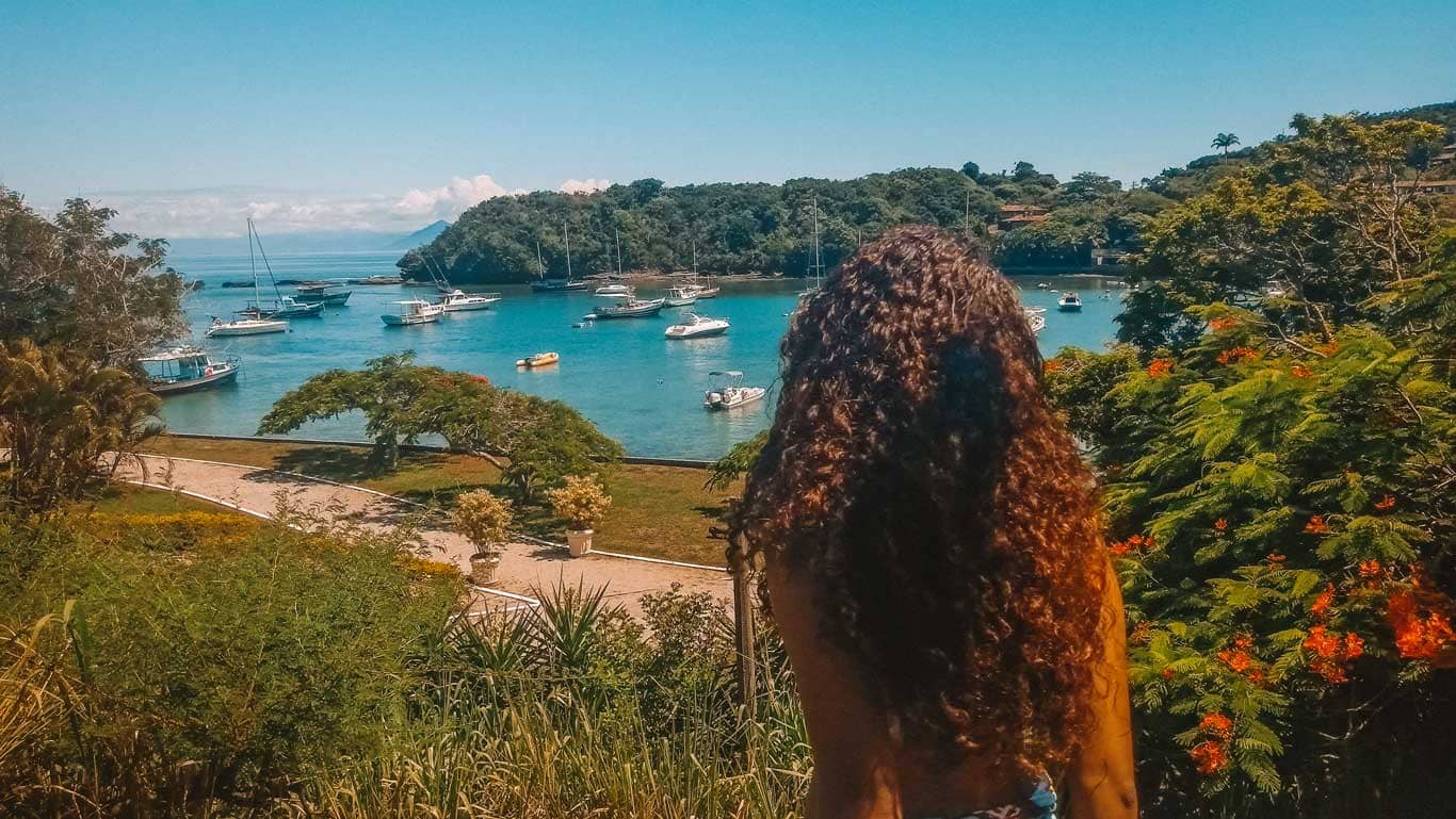 A woman gazing towards the serene Ossos Beach in Buzios, Brazil, with boats anchored in the tranquil blue waters, framed by lush foliage and bright orange blooms under a sunny sky