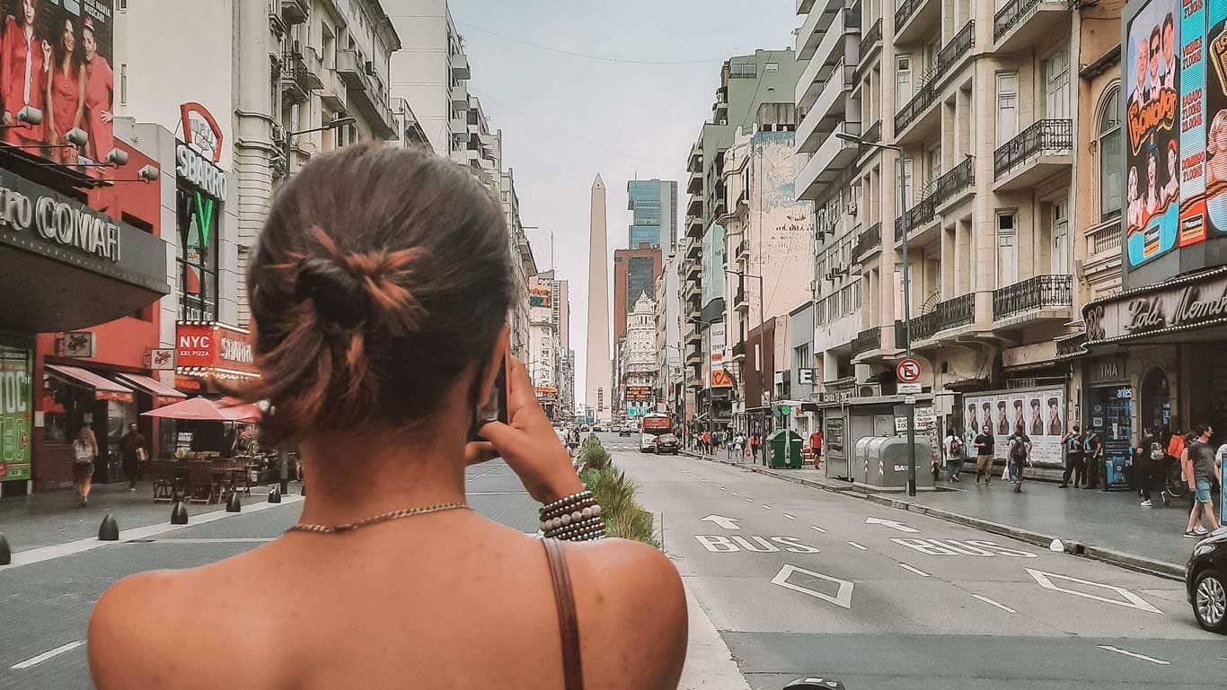 The image captures a bustling urban street scene in Buenos Aires from the perspective of a woman, seen from behind, who is taking a photograph of the street ahead. Notable landmarks such as the Obelisco de Buenos Aires are visible in the distance, framed by an array of street signs, vehicles, and pedestrians. The architecture lining the street reflects a mixture of modern and historical styles, contributing to the dynamic urban atmosphere.