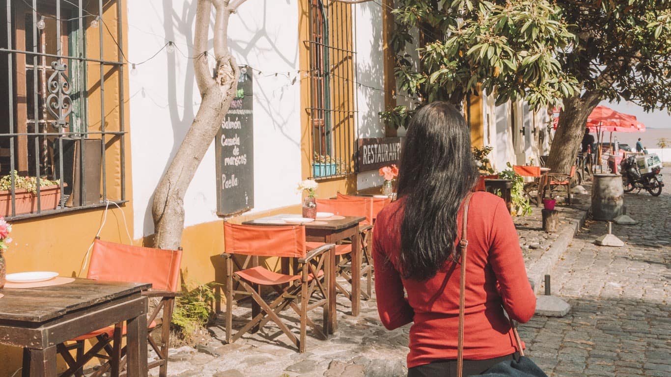 A woman in a red top is seen from behind, walking towards a quaint sidewalk café in Colonia del Sacramento on a sunny day, with rustic wooden tables and red chairs invitingly set out for a relaxing day trip experience.