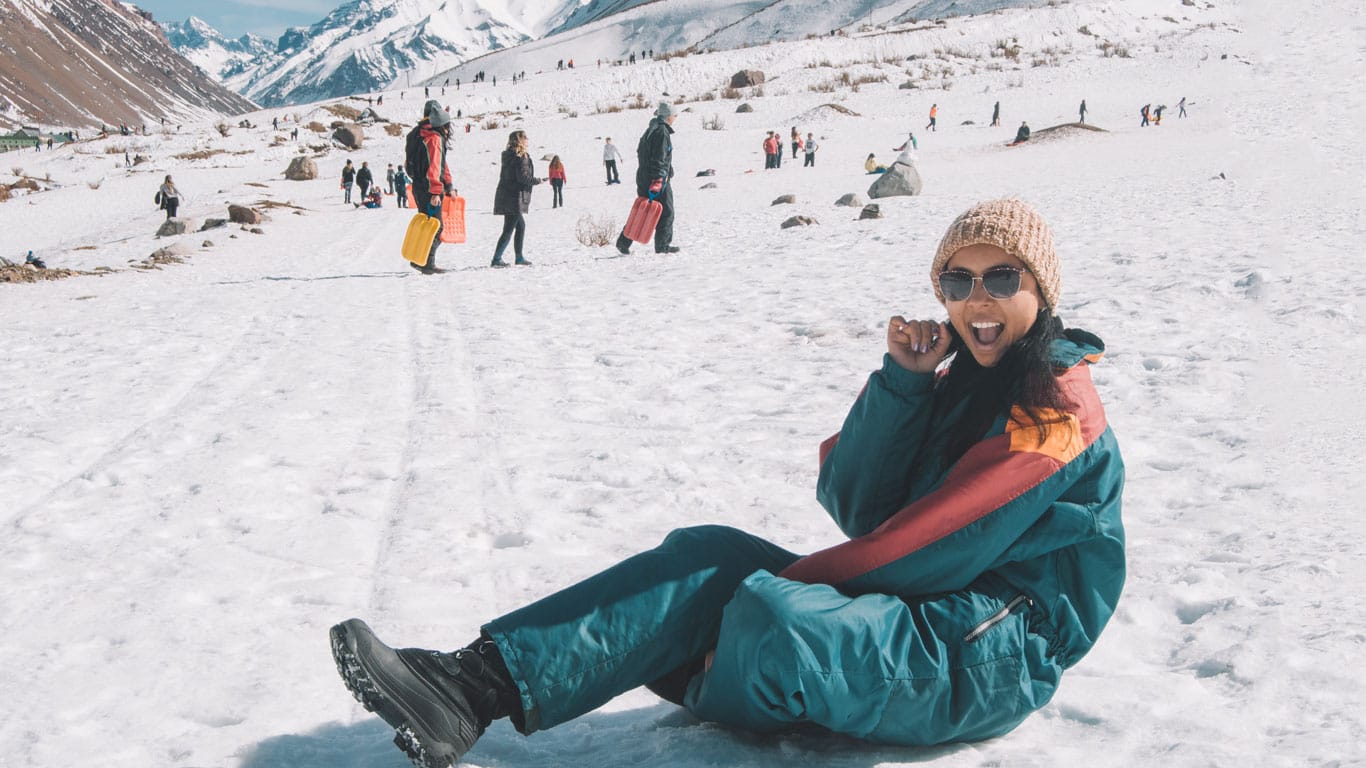 A joyful woman sitting on a snowy slope in Mendoza, Argentina, wearing a colorful winter jacket, sunglasses, and a beige knit hat. She smiles broadly at the camera, enjoying the winter activities around her. The background features a bustling scene of other visitors engaging in snow play, with rugged mountains stretching into the distance.