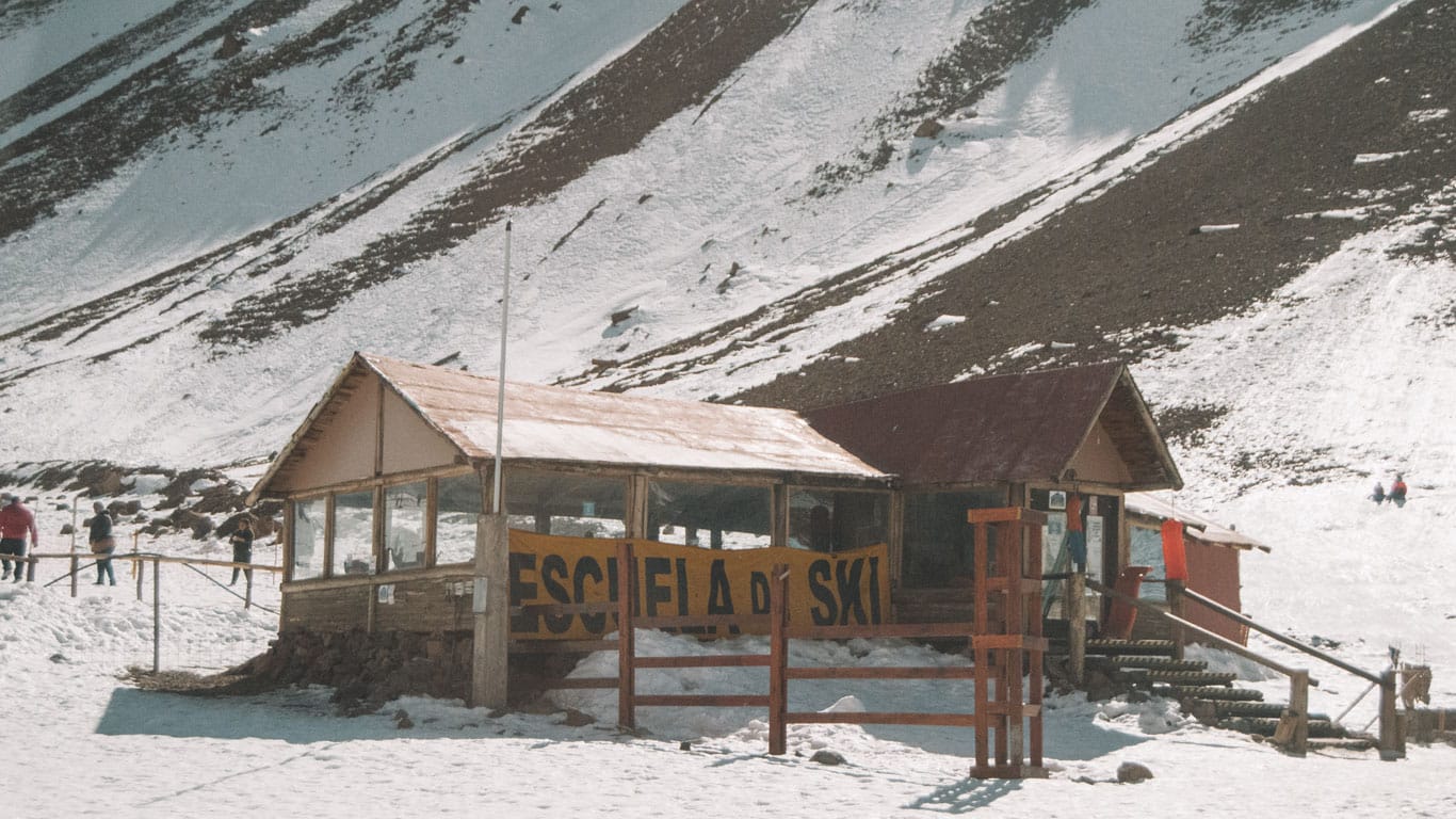 Photograph of a ski school in Mendoza during July. The wooden structure, labeled 'ESCUELA DE SKI', sits at the base of a partially snow-covered mountain. People are visible around the building, enjoying the winter setting under a clear sky.