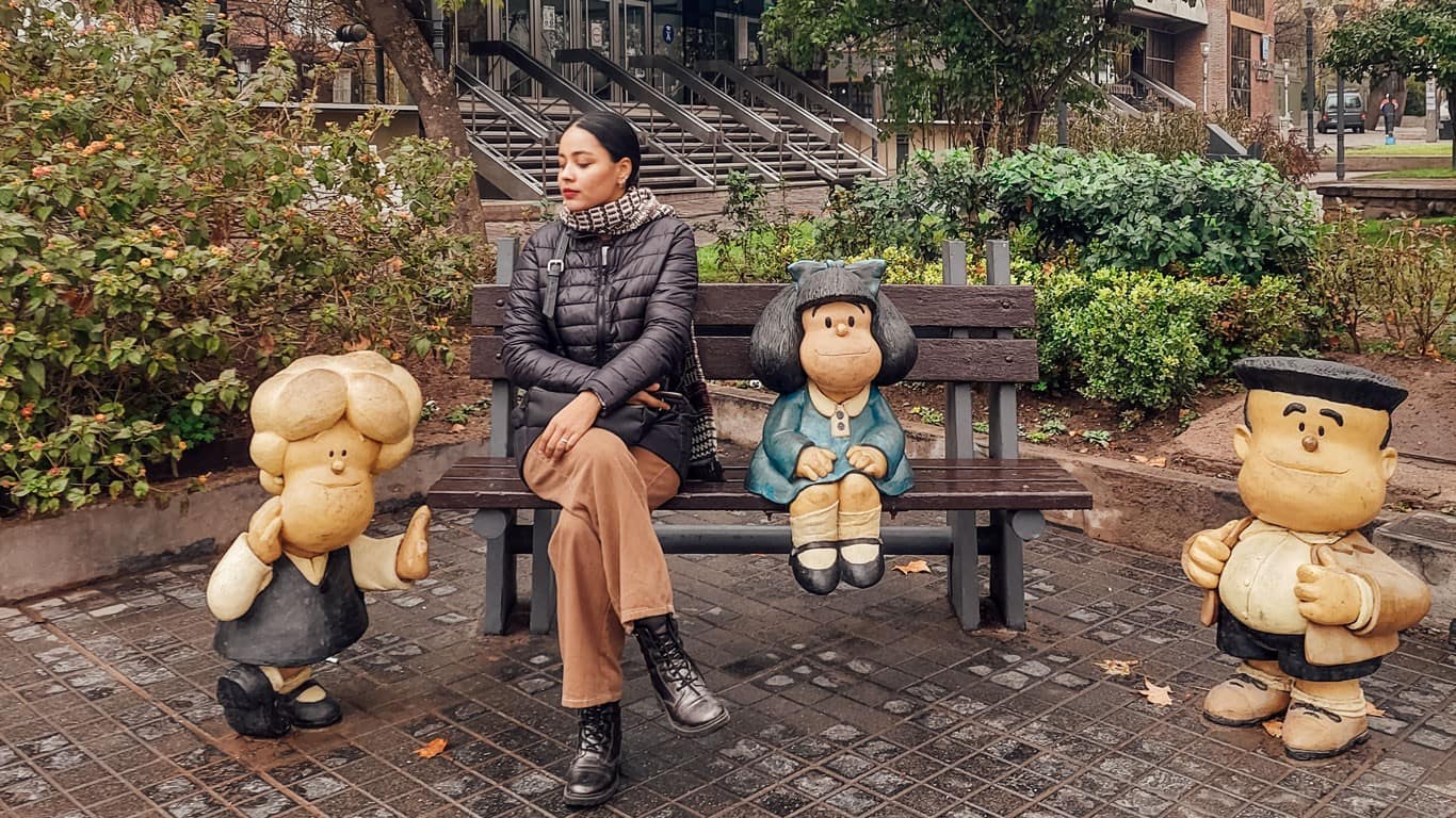 A woman sits on a park bench between colorful cartoon statues in Mendoza, Argentina. On her left is a statue of a young boy with a large hairstyle and oversized shoes, and on her right, a girl with long black hair in a blue dress. Both statues appear cheerful and are styled to look like children's book characters, adding a whimsical touch to the urban setting.