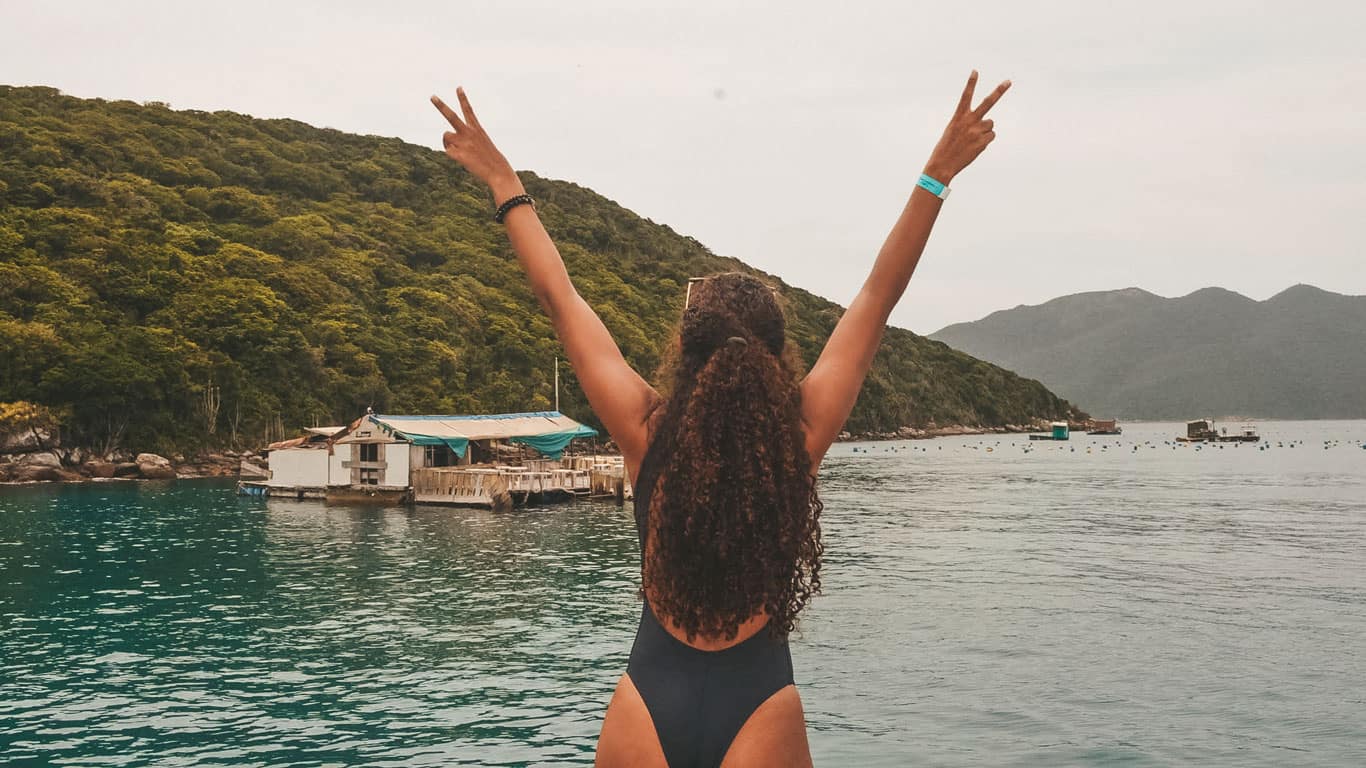 The image shows a woman with her back to the camera, celebrating joyfully with her arms raised and making a peace sign against a picturesque backdrop. She is standing at the waterfront, overlooking a vibrant scene with rustic boats, clear waters, and lush green hills. The atmosphere is vibrant and full of natural beauty, ideal for boat tours in Arraial do Cabo.