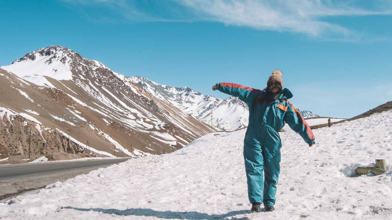 A person in a vibrant orange and blue ski suit joyfully poses with outstretched arms on a snow-covered slope. The backdrop features majestic snow-capped mountains under a clear blue sky, capturing a sunny day in Mendoza in July.
