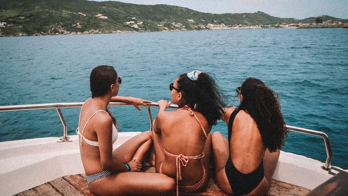 This image captures three women enjoying a scenic boat tour in Arraial do Cabo, Rio de Janeiro. They are seated at the stern of the boat, gazing out at the ocean and the rugged coastline. The women are dressed in swimwear, relaxed and engaged in conversation, against a backdrop of clear blue water and green hills, illustrating a serene day out on the sea.