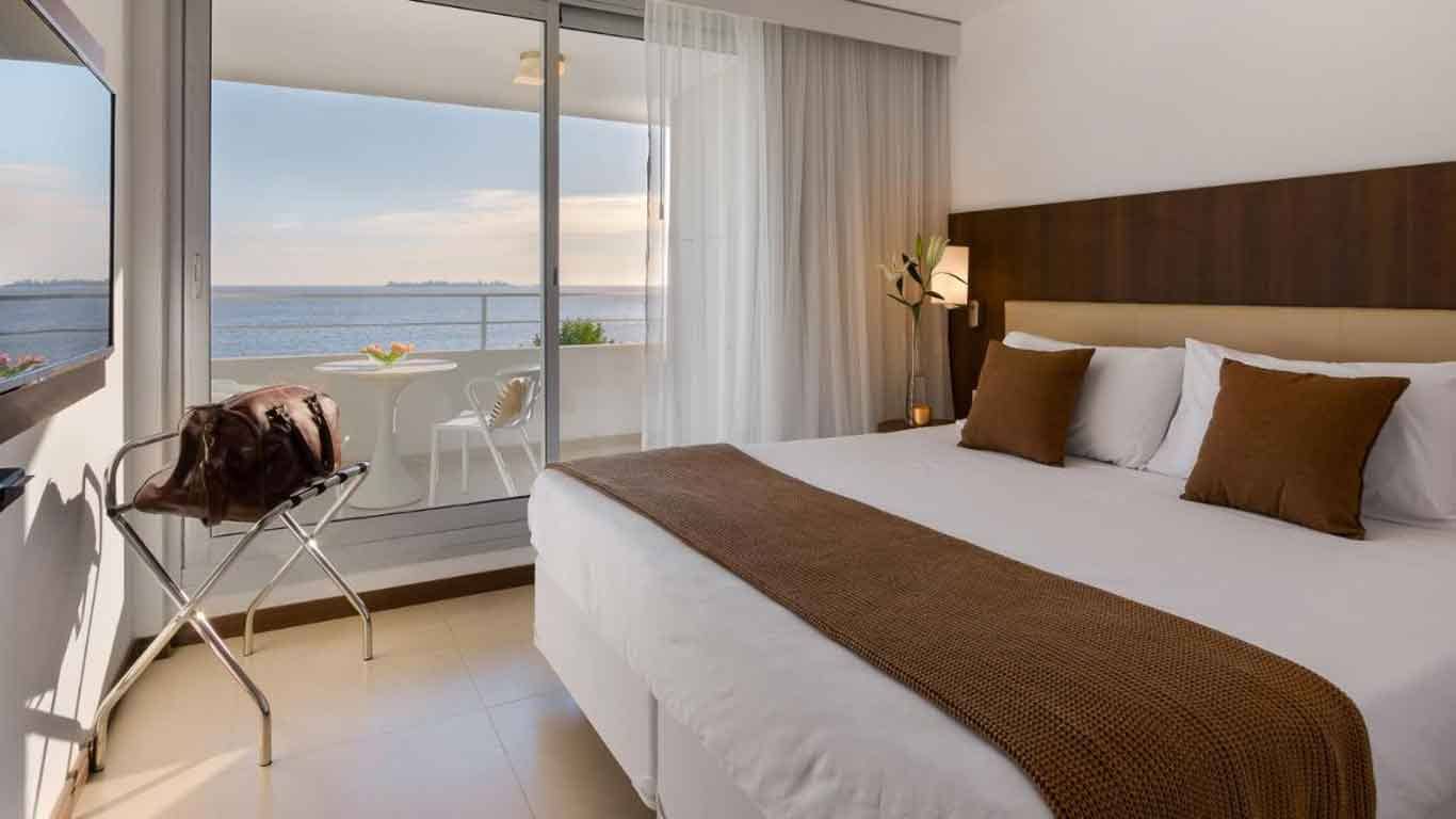 The suite at Dazzler by Wyndham Colonia features a bed with white sheets and a balcony with a stunning view of the Rio de La Plata.