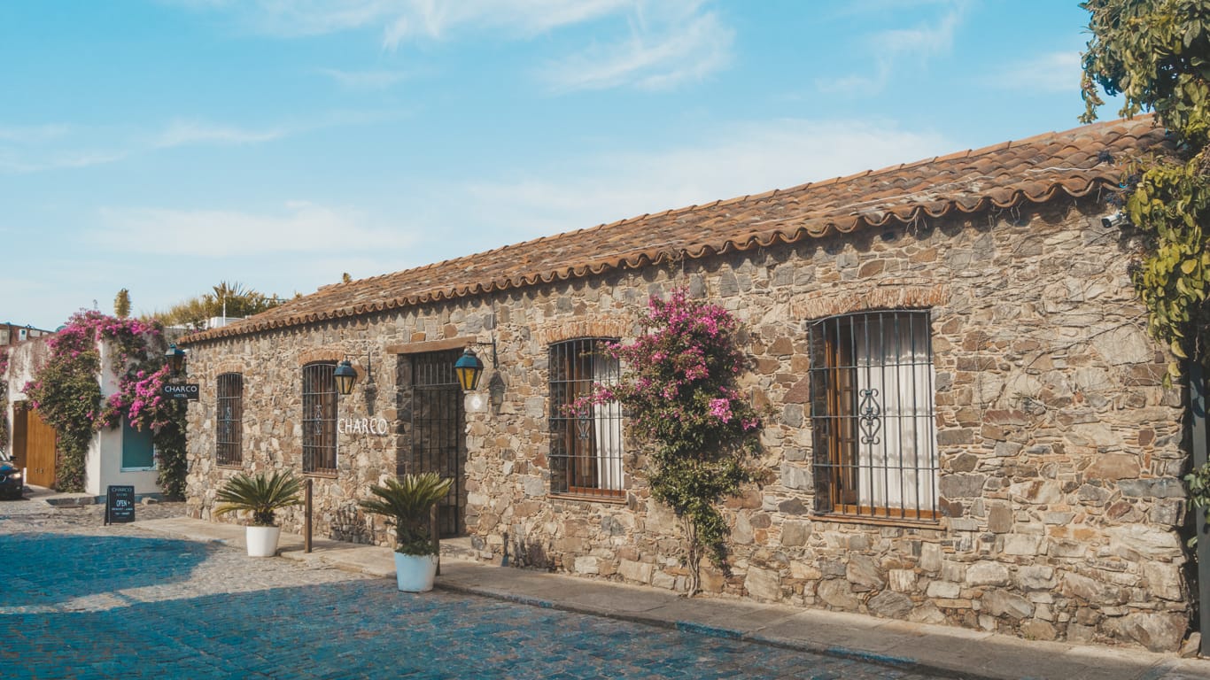 The photo portrays Charco Hotel, a cozy spot with its stone-adorned wall accented by some flowers, situated on a quiet paved street.