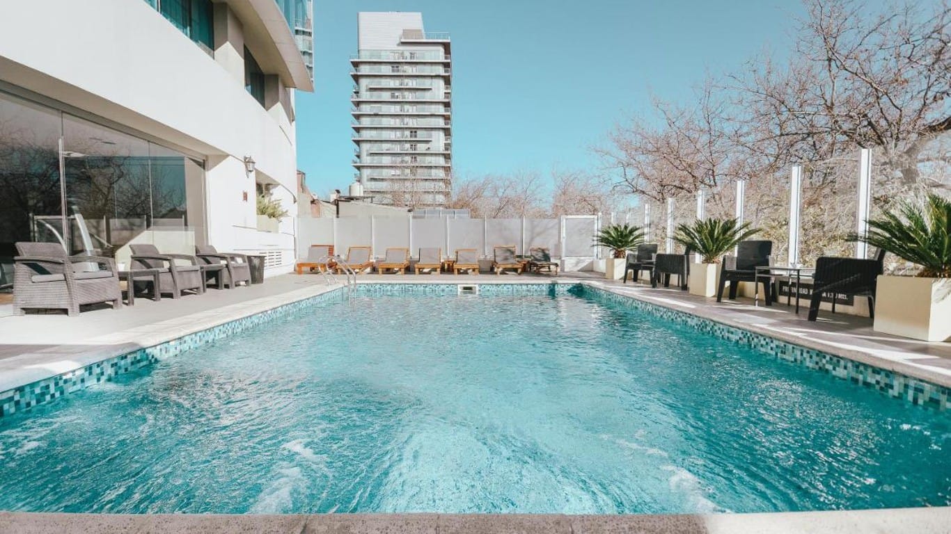 A tranquil outdoor pool area at the Diplomatic Hotel in Mendoza with crystal clear water, surrounded by lounge chairs and palm plants, with the modern hotel building to the left and a clear blue sky overhead.