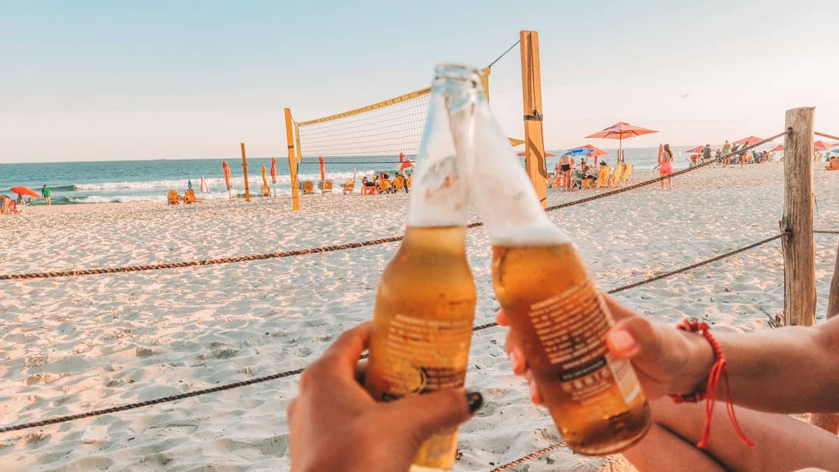 Cheers to beach days! Two hands clinking beer bottles with a volleyball net and vibrant beach umbrellas in the background, capturing the essence of leisure and play at Barra da Tijuca beach in Rio de Janeiro at golden hour.