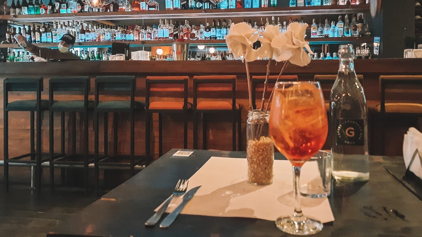 A cozy ambiance at a bar with a table set for one, featuring a refreshing spritz cocktail alongside a bottle of sparkling water, accented by delicate white roses in a vase. The background softly focuses on a bartender behind a well-stocked bar counter.