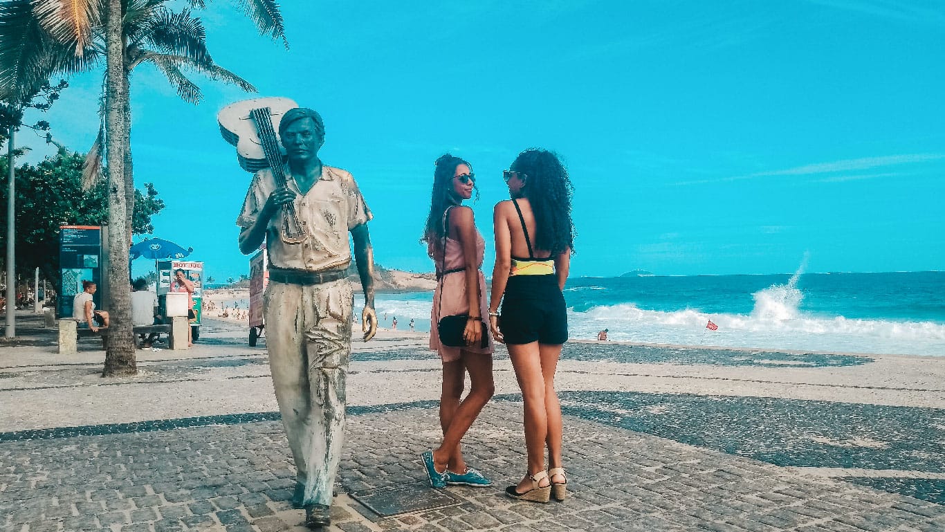 Ipanema Beach comes to life with a bronze statue of a musician greeting visitors, while two people pose for a photo, their relaxed postures mirroring the laid-back vibe of Rio de Janeiro. The palm trees sway gently in the breeze, framing a scenic view of the ocean's powerful waves under the clear Brazilian sky.