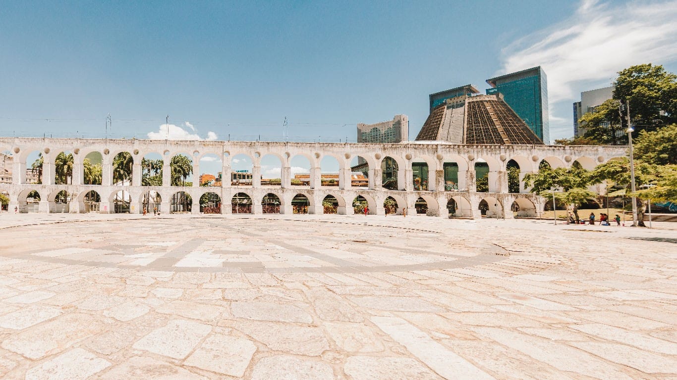 This sunlit view showcases the elegant colonial structure of the arches contrasting with the modern skyscrapers in the background, while the spacious foreground of the square invites residents and tourists to explore the Lapa region, an incredible option of where to stay in Rio de Janeiro.