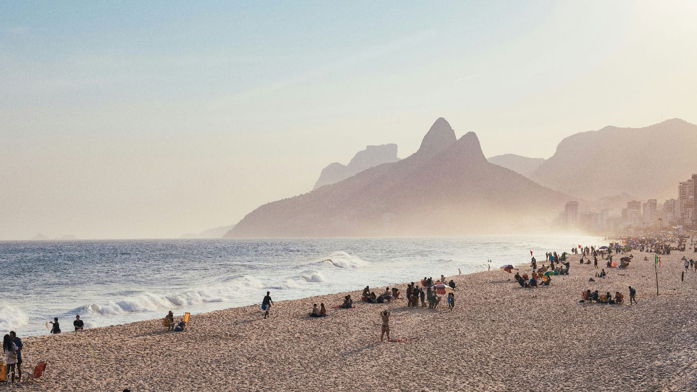 Leblon Beach in Rio de Janeiro is bathed in a golden haze as the day winds down, with the iconic Two Brothers Mountain standing majestically in the background. Beachgoers enjoy the lingering warmth of the sun, dotting the expansive sandy shore that stretches towards the urban silhouette, creating a serene and inviting atmosphere for relaxation and socializing.
