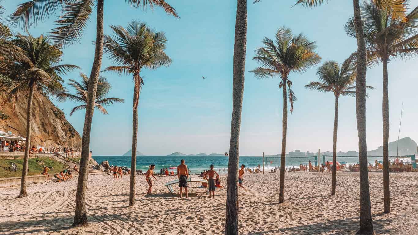 The picturesque Leme Beach in Rio de Janeiro is framed by towering palm trees, with a lively beach scene of sunbathers and volleyball players enjoying the golden sands and clear skies.