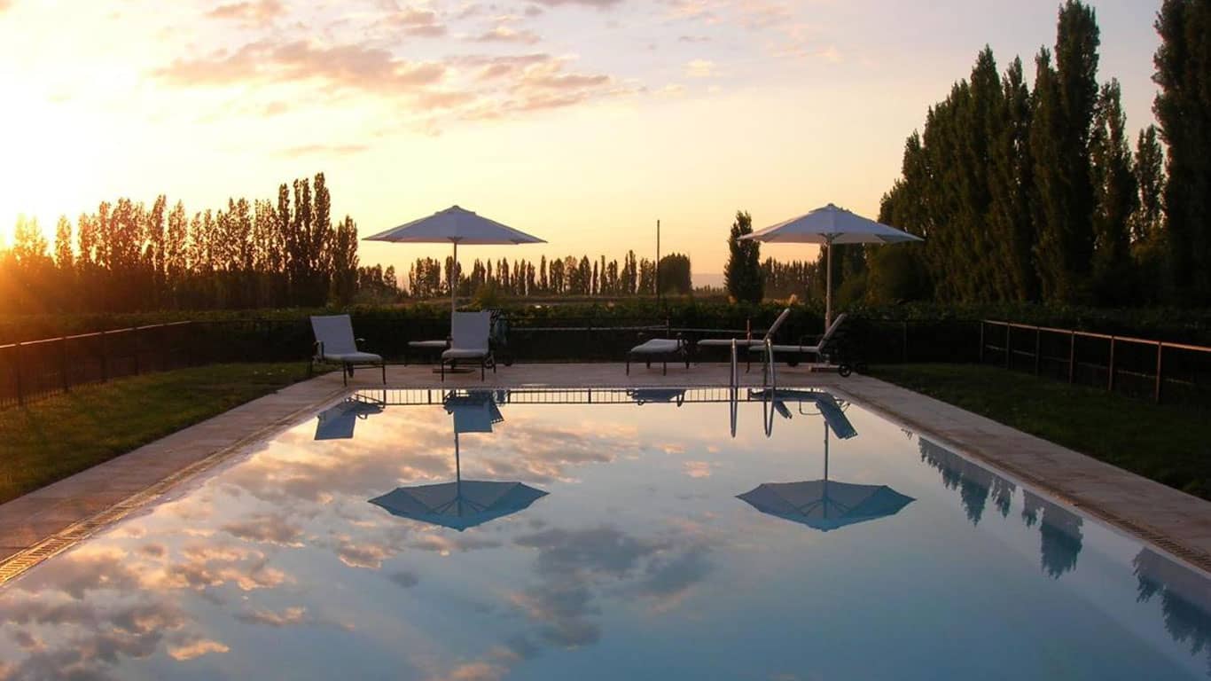 Dusk settles over the serene Posada Salentein pool in Mendoza, reflecting the soft golden sky and white clouds above, with the silhouette of tall trees in the background and poolside lounge chairs inviting relaxation.